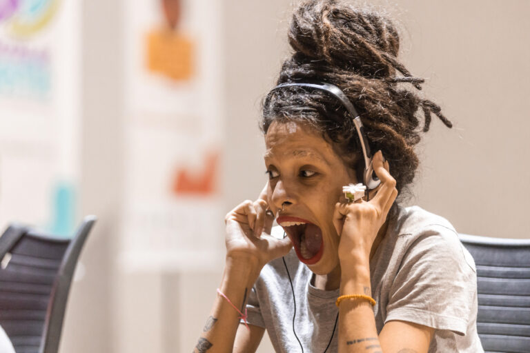 Descrição da imagem: Foto colorida. O rosto de uma mulher, que usa os fones da audiodescrição, tem a boca bem aberta, as sobrancelhas arqueadas, e expressa alegria e surpresa. Suas mãos seguram os fones junto ao ouvido. Ela tem o cabelo castanho, comprido, com dread, preso no alto da cabeça, olhos também castanhos, usa piercing, estilo ferradura, no septo. Anel de flor branca no dedo médio da mão esquerda e camiseta de malha cinza.Tem tatuagens em ambos os braços.